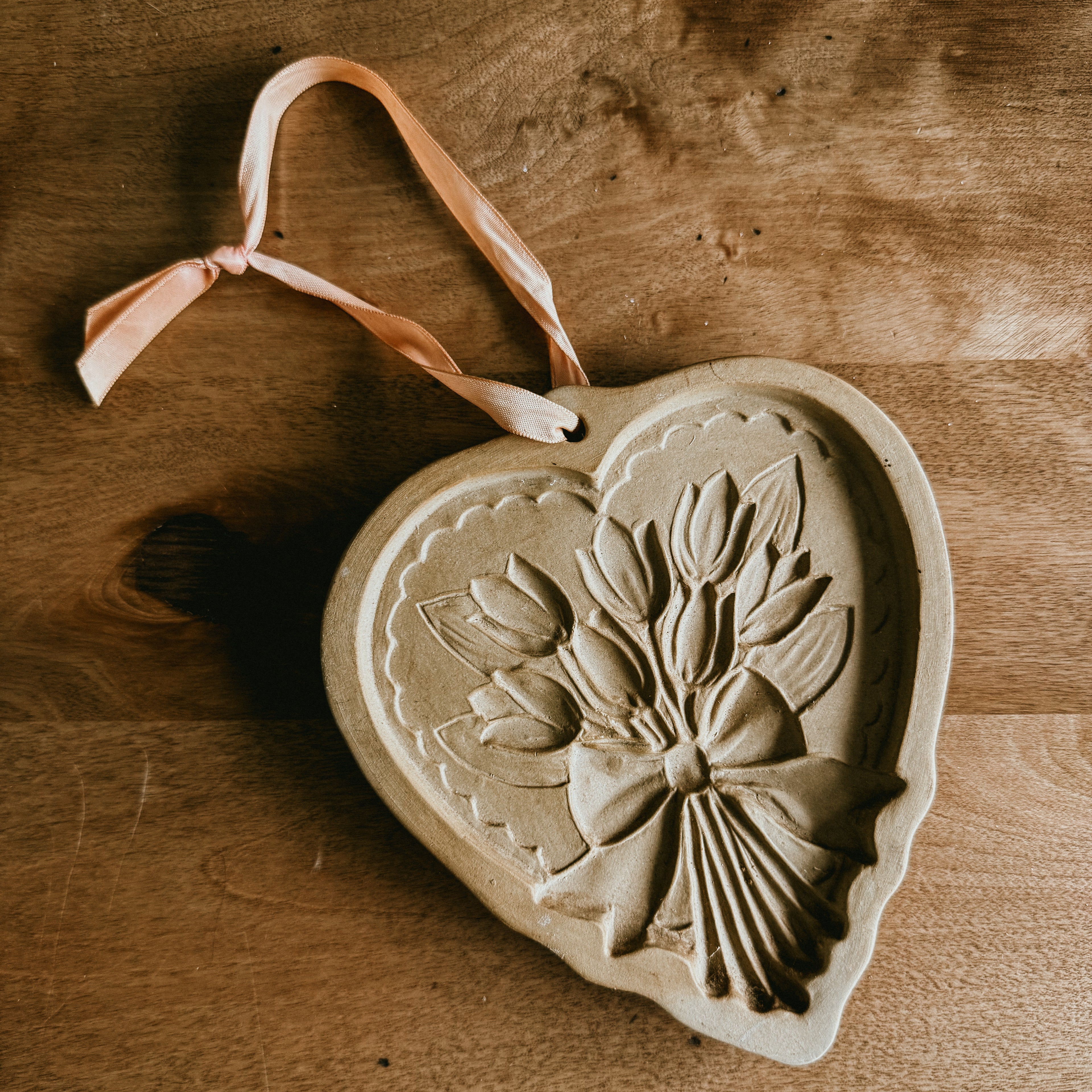 A tan-colored, heart-shaped stoneware cookie mold featuring a detailed relief of a tulip bouquet tied with a bow, resting on a wooden surface with a pink ribbon for hanging.
