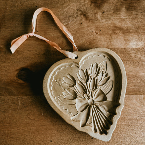 A tan-colored, heart-shaped stoneware cookie mold featuring a detailed relief of a tulip bouquet tied with a bow, resting on a wooden surface with a pink ribbon for hanging.