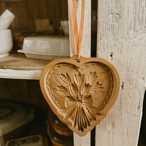 A tan-colored, heart-shaped stoneware cookie mold featuring a detailed relief of a tulip bouquet tied with a bow, resting on a wooden surface with a pink ribbon for hanging.