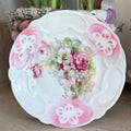 Antique German porcelain dessert plate featuring a central bouquet of pink roses and white lilies with a scalloped edge and pink airbrushed borders.