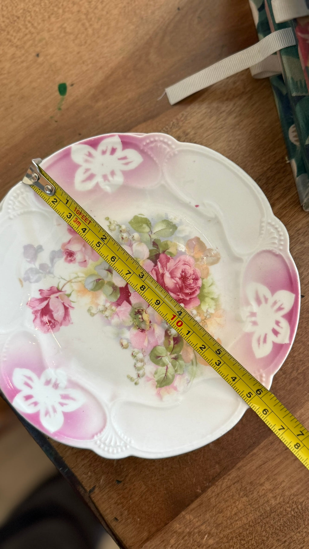 A yellow measuring tape laid across a 6.5 inch antique German porcelain floral plate to show exact diameter sizing.