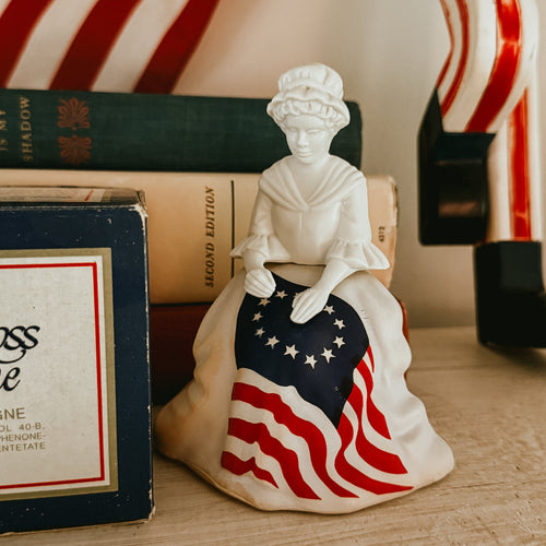 Vintage 1976 Avon Betsy Ross milk glass figurine cologne bottle standing next to its original blue illustrated retail box.